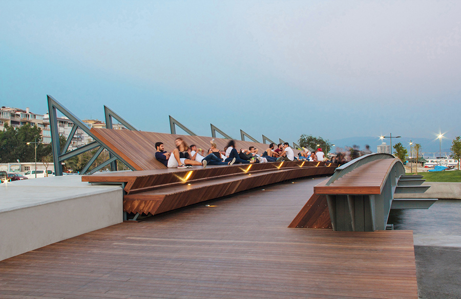 Bostanlı Footbridge: A Symbol of Connectivity and Leisure Studio Evren Başbuğ Architects envisioned Bostanlı Footbridge as a vital component of the 'Karşıkıyı' concept within the 'İzmirSea' coastal regeneration project. This architectural marvel serves not only as a functional connector but also as a captivating leisure spot. The footbridge spans Bostanlı Creek, seamlessly linking both sides and completing the continuous coastal promenade. Its unique design, featuring a slightly bow-shaped longitudinal section and specially crafted girder geometry, facilitates the passage of small boats underneath. Additionally, the bridge grants access to a floating pontoon nestled within the creek. Designed with the site's distinct characteristics in mind, the footbridge offers a picturesque view of the bay on one side and the city on the other. Its asymmetrical cross-section, crafted from cascading thermo-wood surfaces on a steel frame, invites users to pause and admire the scenery. Rather than being a mere passageway, this structure transcends its infrastructural role to become a public leisure destination integrated with its surroundings. Bostanlı Sunset Lounge: Embracing Nature's Beauty Situated on a west-facing coastal fragment in Karşıyaka, Bostanlı Sunset Lounge complements the footbridge with its inviting design. Consisting of cascading thermo-wood covered platforms, this urban oasis creates a serene space where visitors can bask in the beauty of the setting sun and the sea. The lounge's simplicity in surface geometry fosters a direct connection with nature, encouraging visitors to immerse themselves fully in the tranquil ambiance. Like its counterpart, the footbridge, the Sunset Lounge evokes a sense of warmth and welcome, largely influenced by the natural texture of the materials used. By reviving the tradition of sunset watching—a cherished ritual in İzmir's urban memory—the Sunset Lounge fosters community engagement and shared experiences among city residents. Embodying Urban Renewal and Community Engagement Together, Bostanlı Footbridge and Sunset Lounge epitomize the 'Karşıkıyı' proposal's vision for the Karşıyaka coastline. These installations, characterized by their natural, inclusive design, serve as 'counter-spaces,' offering respite from the bustling urban environment. Rooted in the social, geographical, and historical context of their surroundings, these structures invite visitors to embrace the concept of 'idleness' and indulge in moments of relaxation. Furthermore, they align seamlessly with İzmir's overarching vision of an 'Easy Way of Living,' as outlined by the 'İzmirSea' coastal regeneration project. In essence, Bostanlı Footbridge and Sunset Lounge not only enhance connectivity and accessibility along the coastline but also foster a sense of community and appreciation for İzmir's natural beauty.